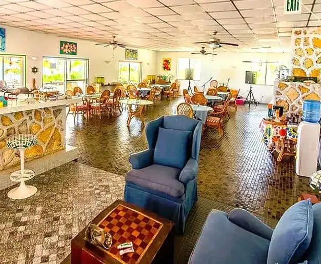 dining-img3 | Greenwood Beach Resort, Cat Island, Bahamas Spacious indoor lounge with blue armchairs, a chessboard table, round tables and wicker chairs, stone fireplace, and ceiling fans.