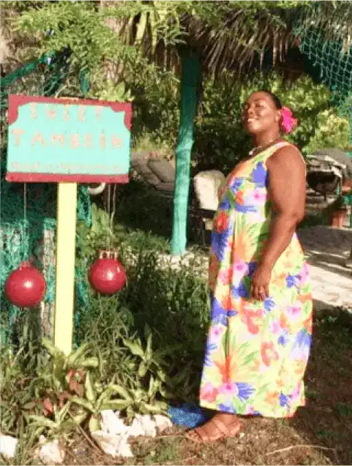 discover-bush-medicine | Greenwood Beach Resort, Cat Island, Bahamas Woman in a bright floral dress stands beside a turquoise sign in a tropical garden, with red decorative balls nearby.