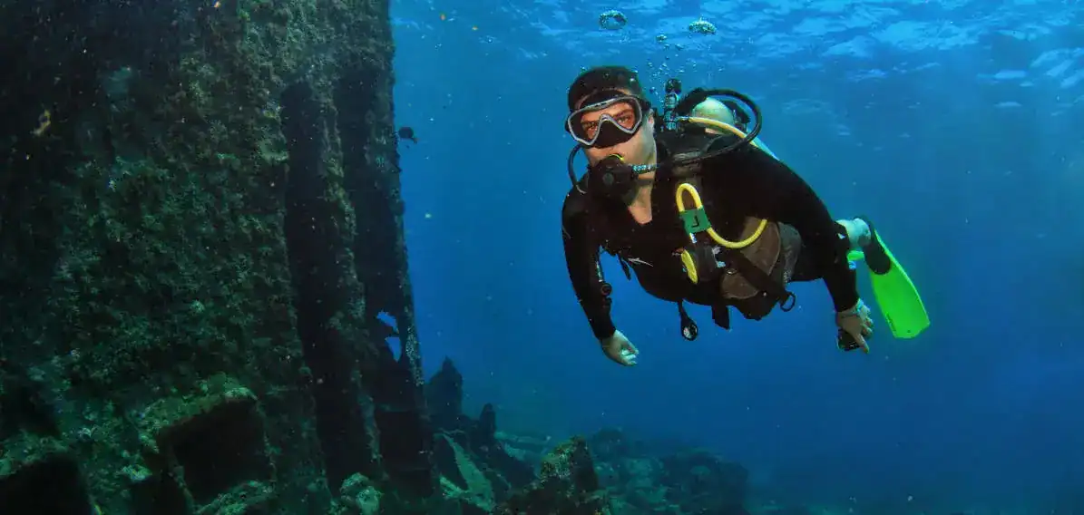 scuba | Greenwood Beach Resort, Cat Island, Bahamas Scuba diver in a black wetsuit and mask gliding underwater near a rocky reef wall, blue open water around him